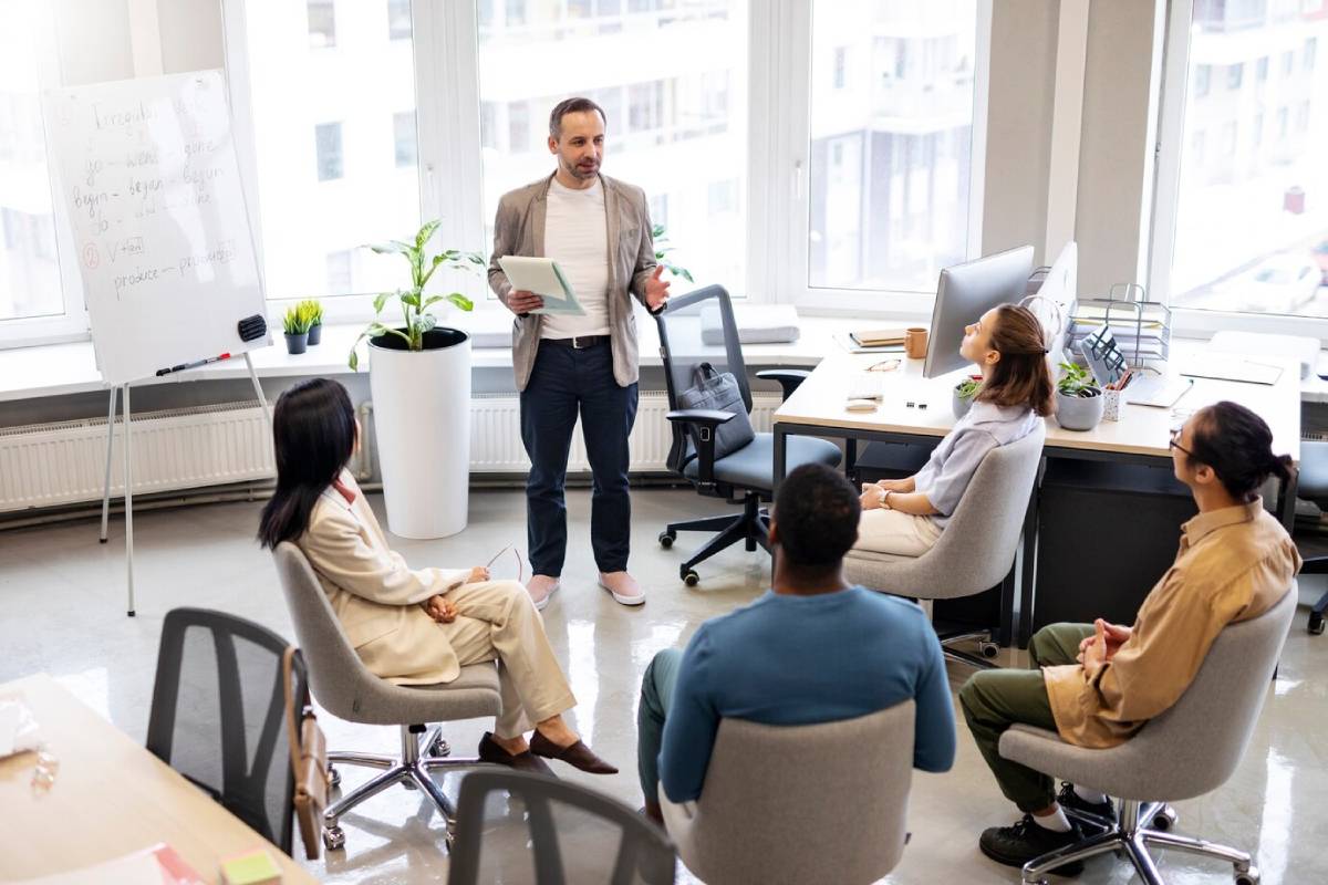 A man presents to coworkers during a meeting in an office with large windows. 