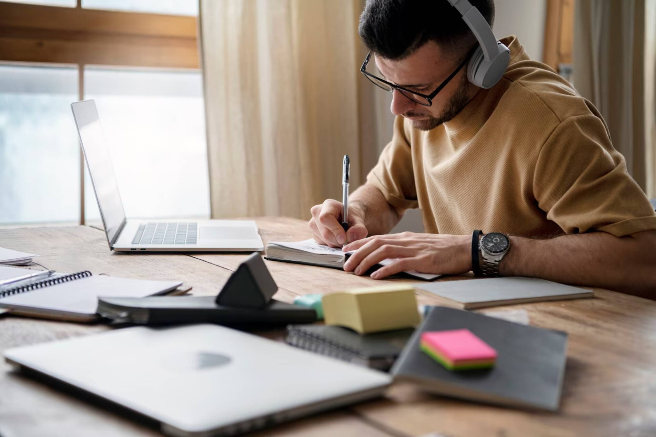 Man with headphones writing notes at a desk with a laptop and stationery. 