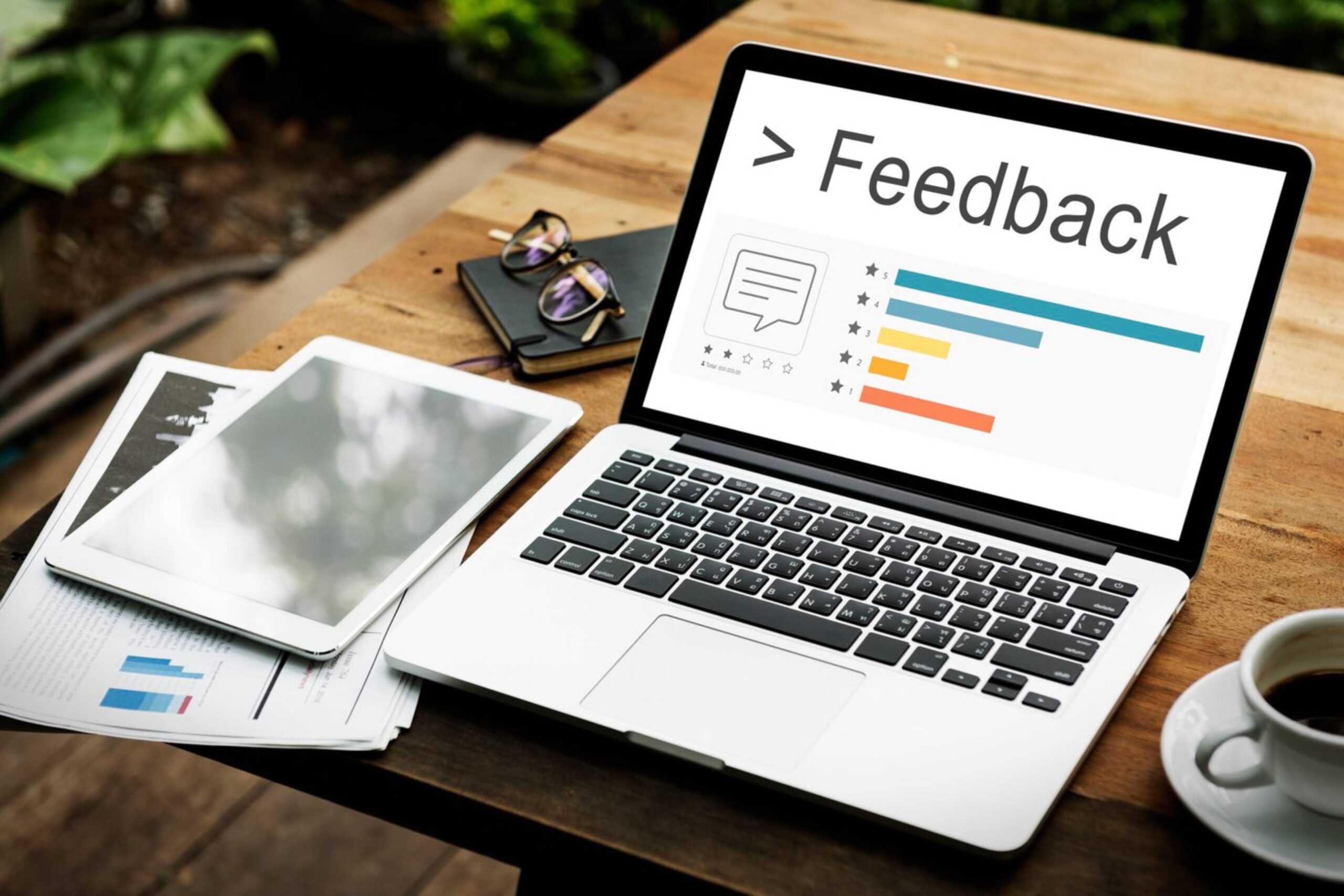 A laptop displaying a feedback screen, surrounded by papers, a coffee cup, and glasses on a wooden table outdoors. 