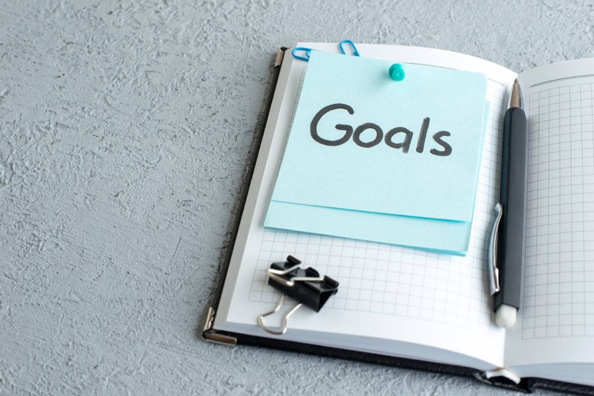  A notepad with a 'Goals' sticker, clipped papers, and a pen on a textured grey surface.