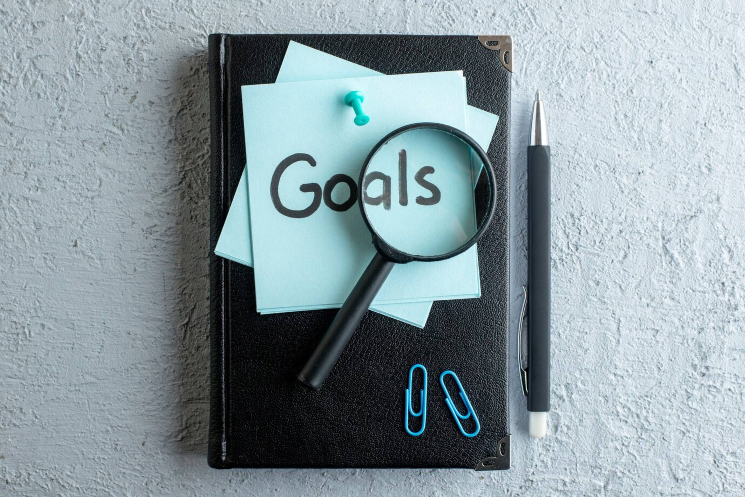  A magnifying glass and pen on a black notebook with a 'Goals' sticky note and paper clips on a textured surface.