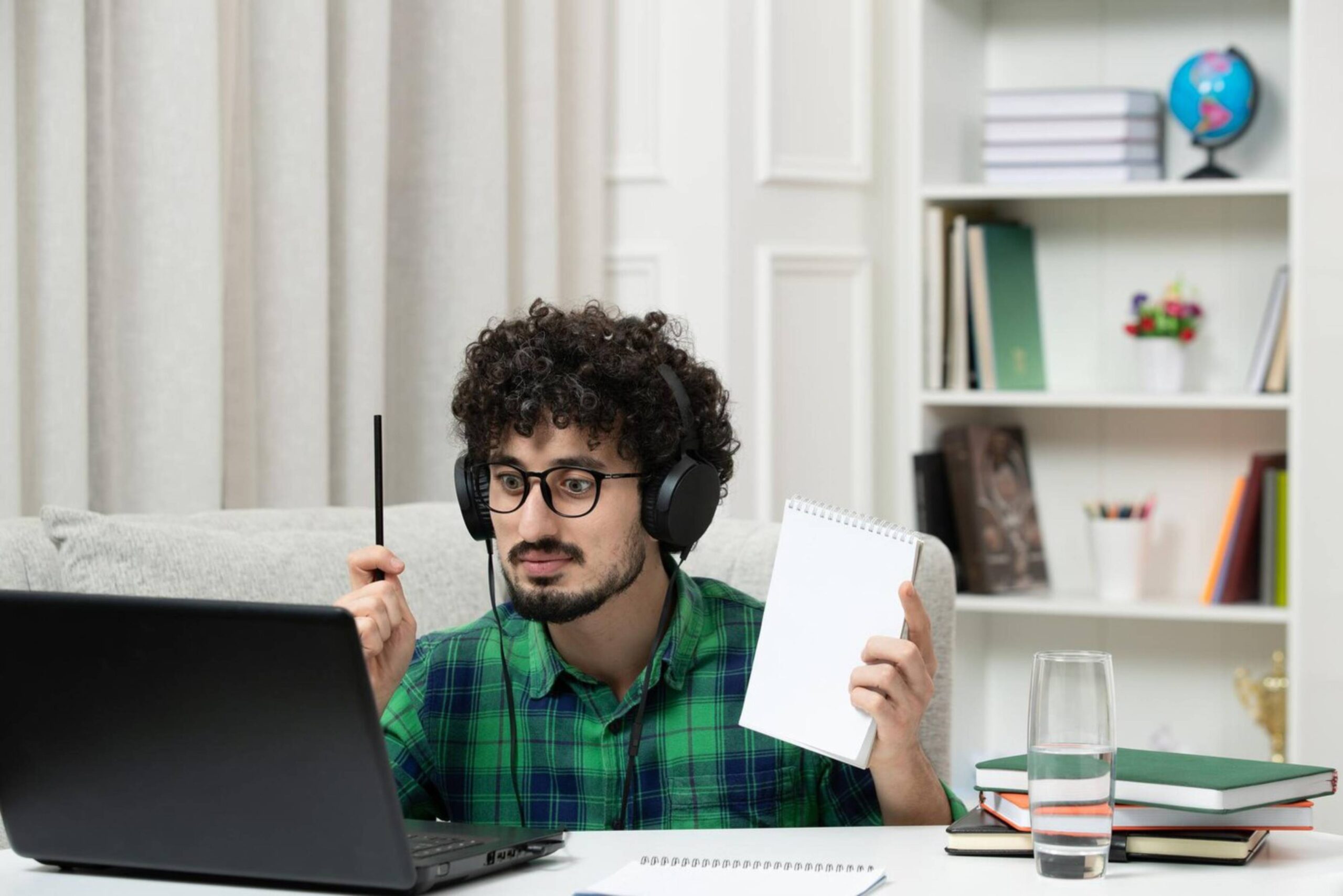 Man with headphones sits at a desk, holding a pen and notepad, interacting with a laptop at home. 