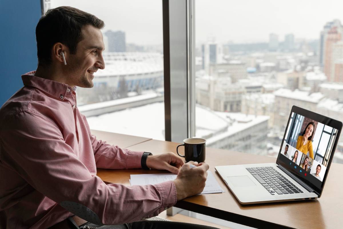 Man in pink shirt smiles during a virtual meeting with colleagues on laptop in a snow-view office. 