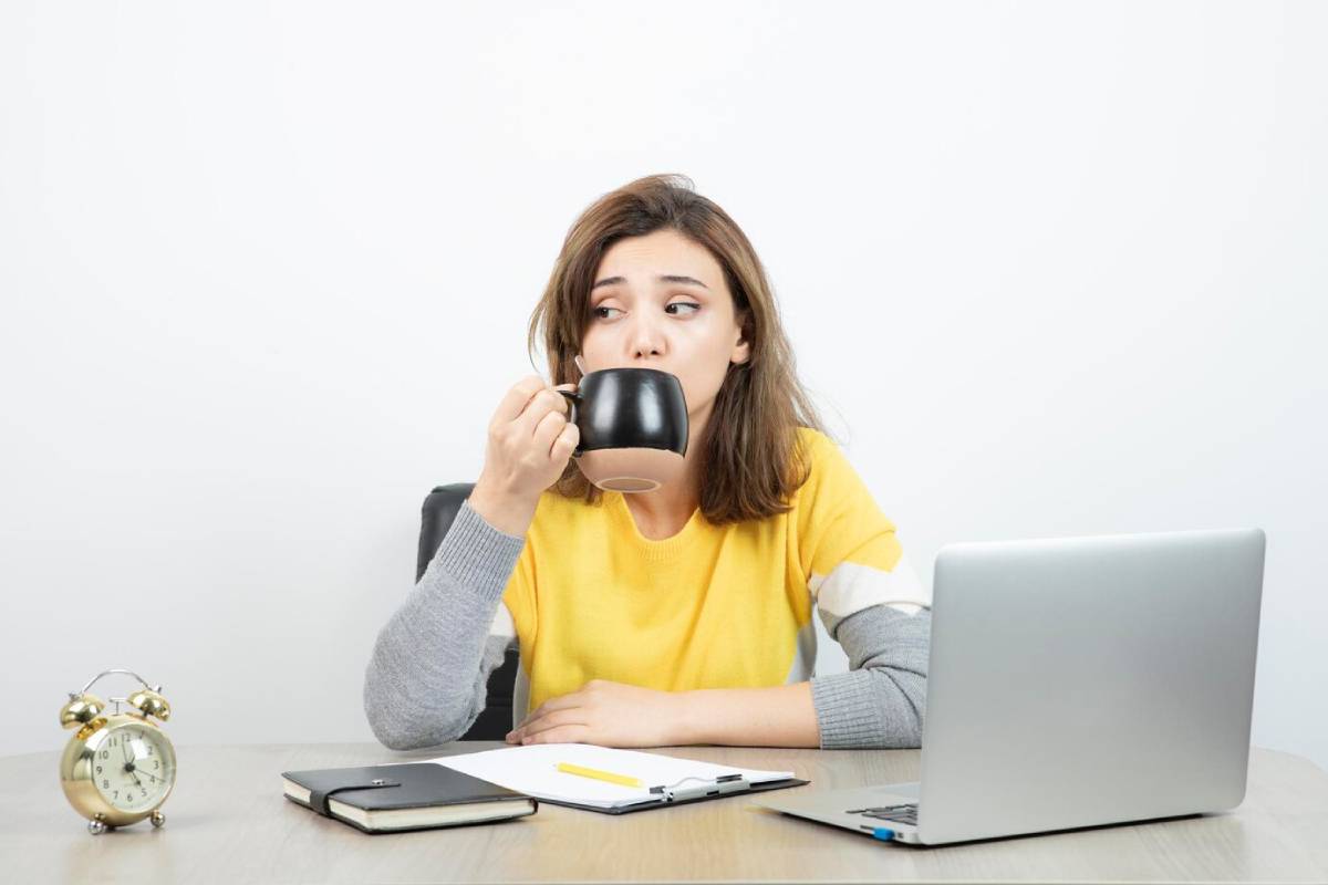 Young woman drinking coffee while working on a laptop, with a clock and notepad on the desk. 