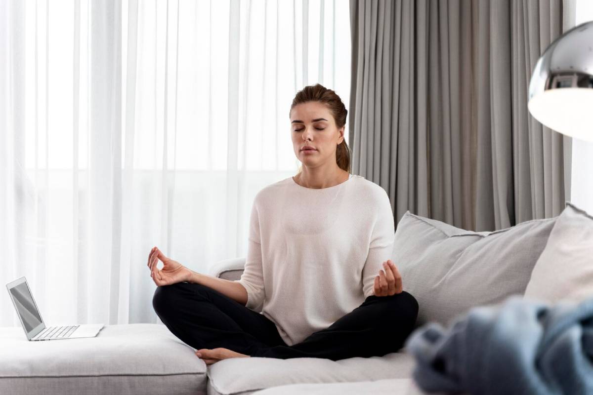  Young woman meditating on a sofa in a bright living room, with a laptop beside her.