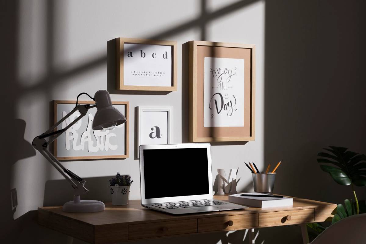  A modern workspace with a laptop, desk lamp, framed typographic art, and houseplants, bathed in natural sunlight. 