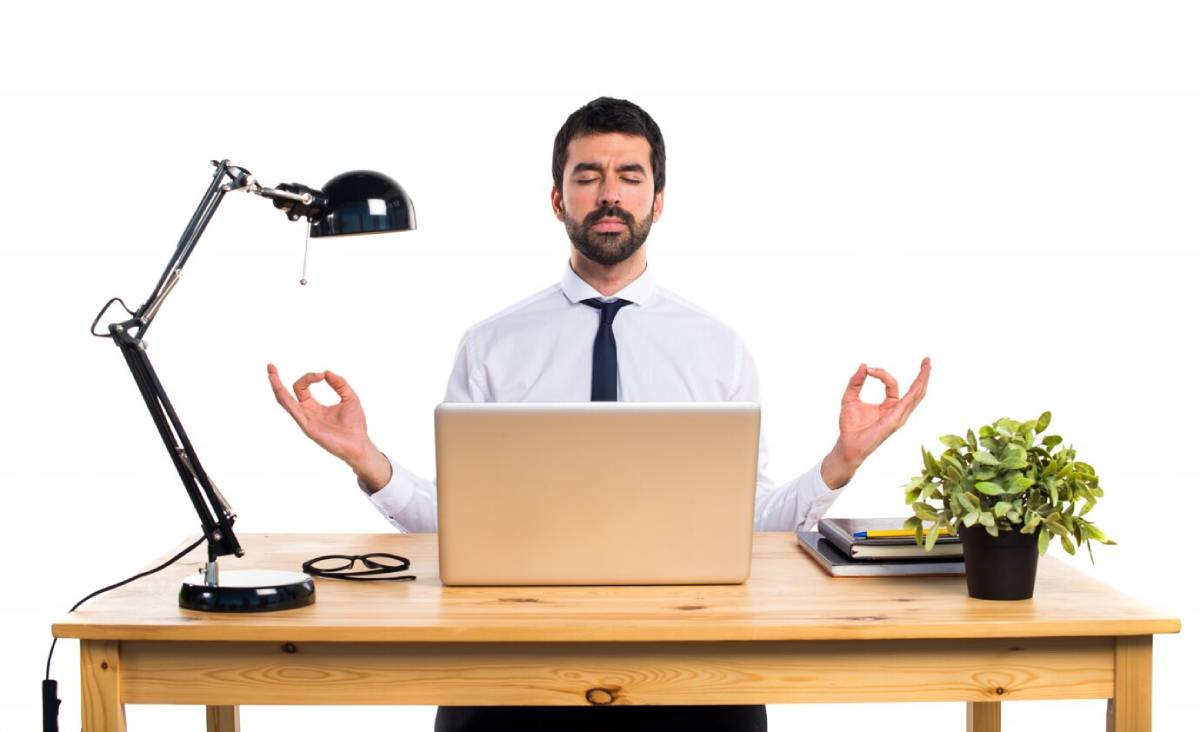  Man in business attire meditating at his office desk with laptop and desk lamp, isolated on a white background.