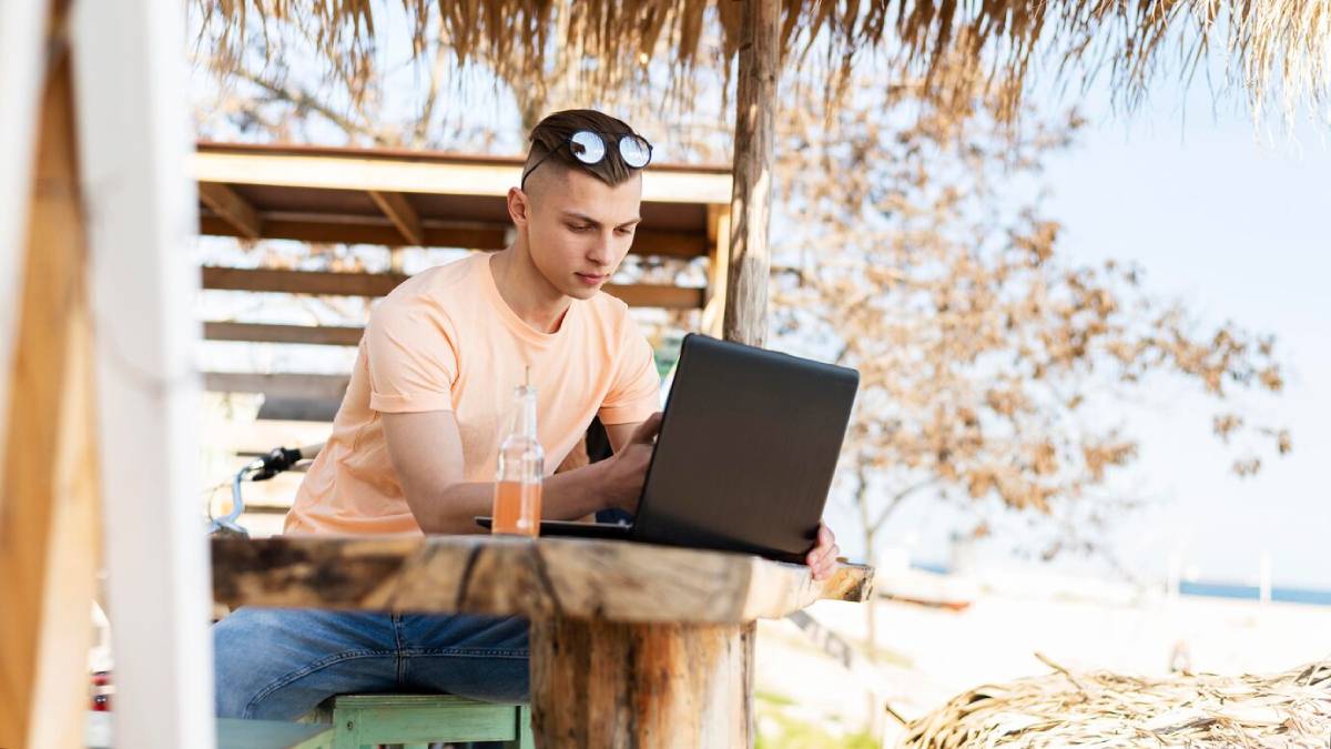  Young man using laptop at a beach hut with a drink beside him.