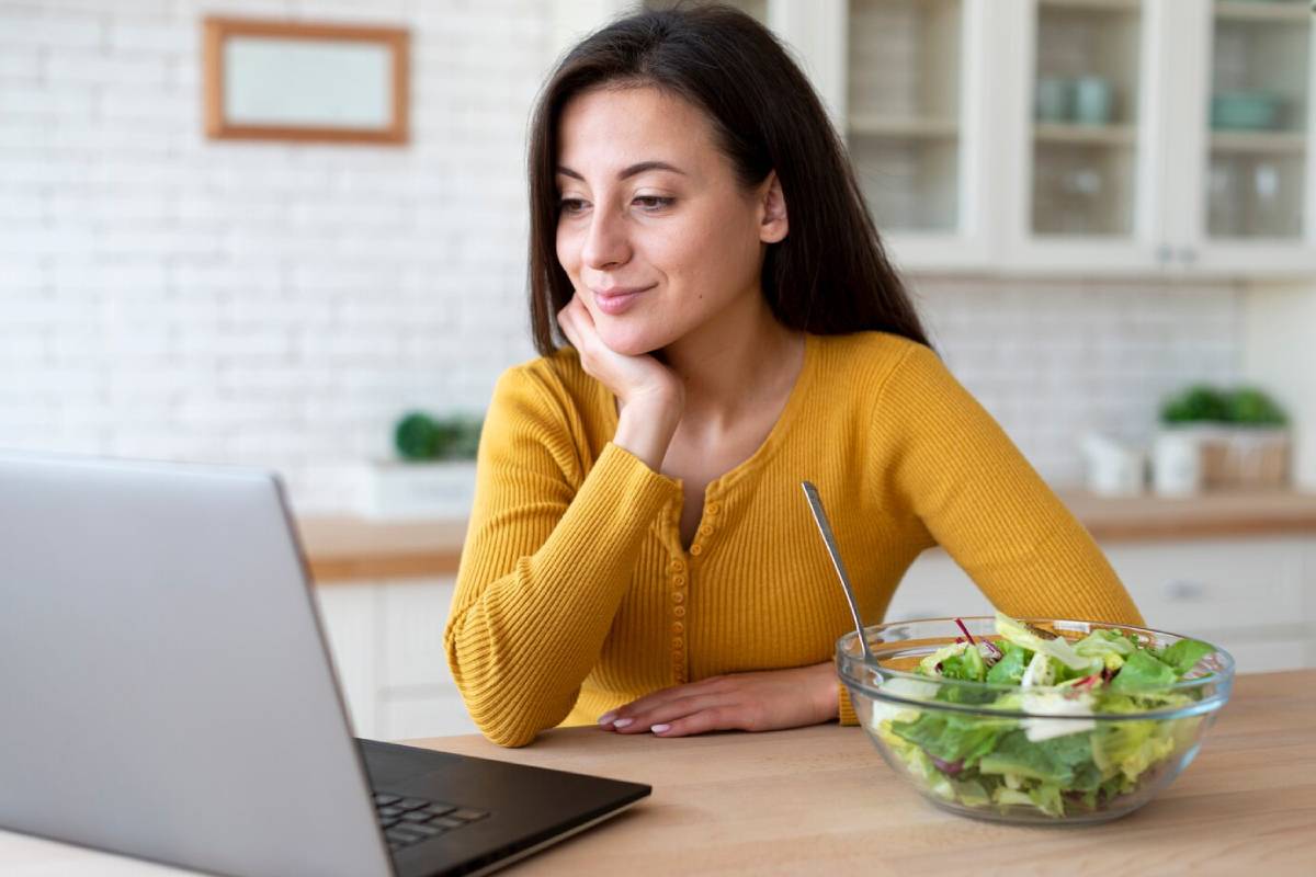  A woman in a yellow sweater smiles while looking at her laptop beside a bowl of salad in a bright kitchen.