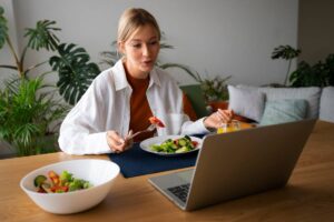 A woman eating a salad while working on her laptop at a dining table.